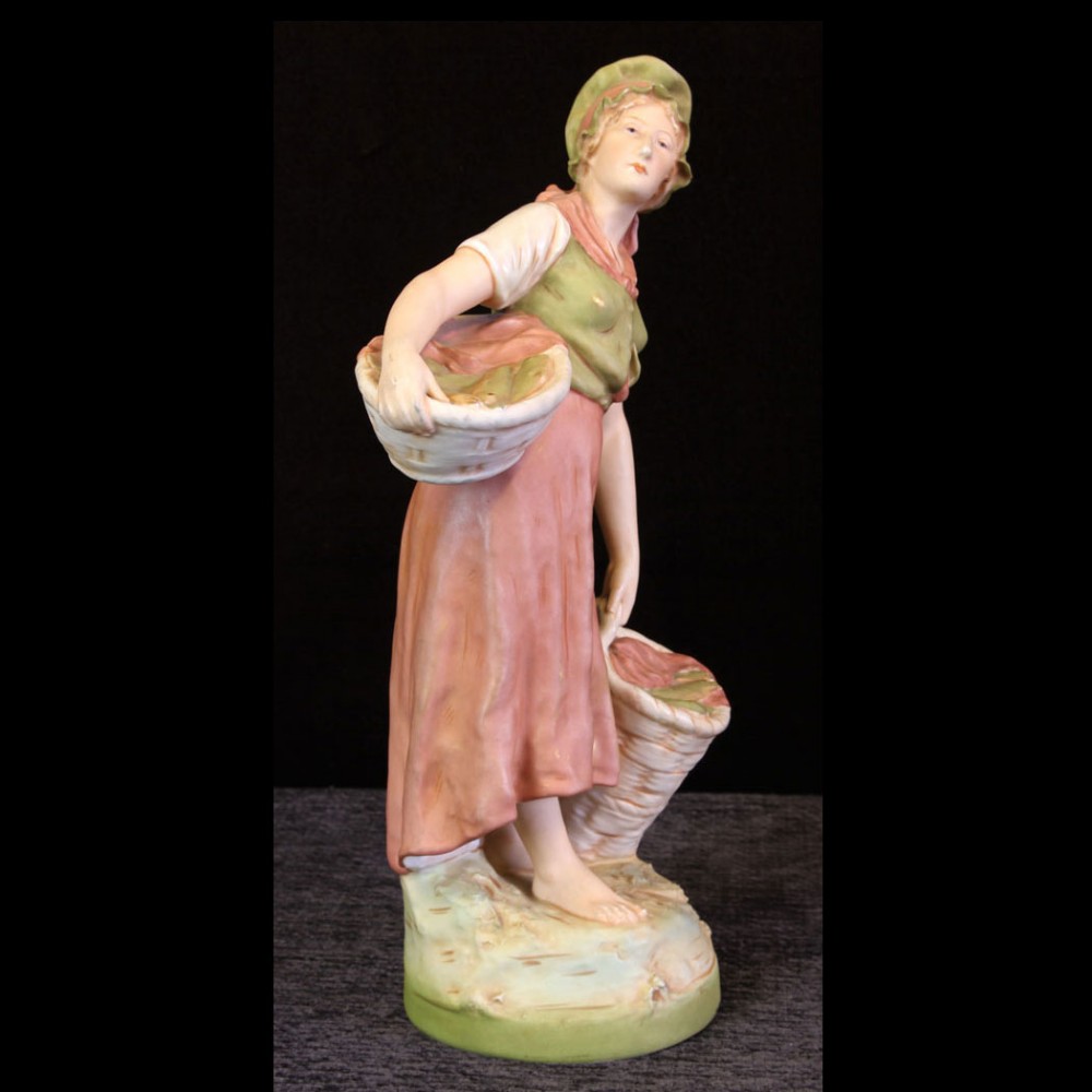 An antique Royal Dux porcelain figure depicting a young girl in a pastel pink dress and green bonnet carrying two woven baskets.