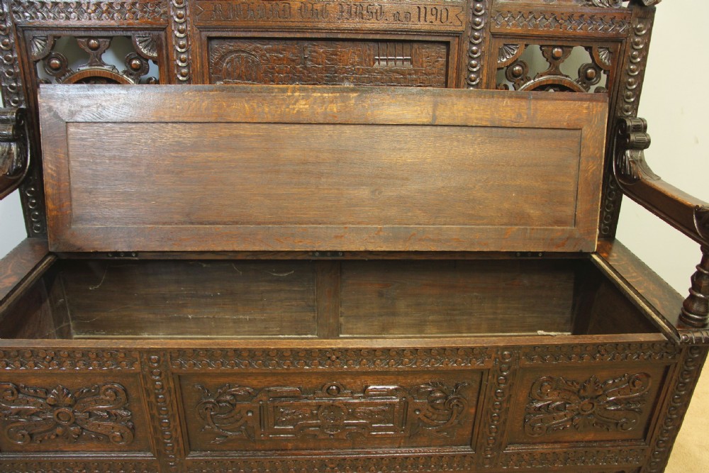 A dark oak Victorian settle with relief-carved pictorial panels, carved mask armrests, wheel motif backrest panels, and a hinged storage seat. View IX