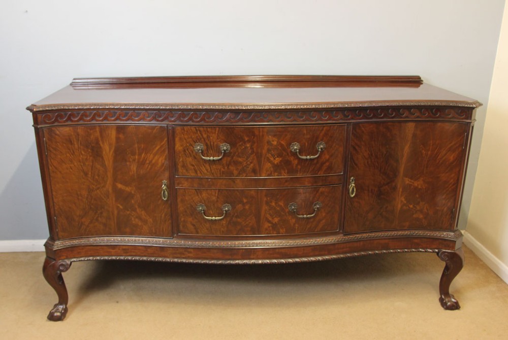 An antique dark mahogany serpentine sideboard featuring central drawers, flanking cupboard doors, brass drop handles, carved frieze moulding, and cabriole legs.