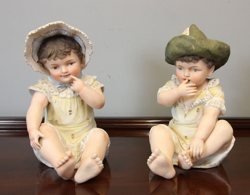 A pair of antique bisque porcelain piano baby figurines depicting seated infants wearing bonnets and lace-trimmed garments with gilded accents.
