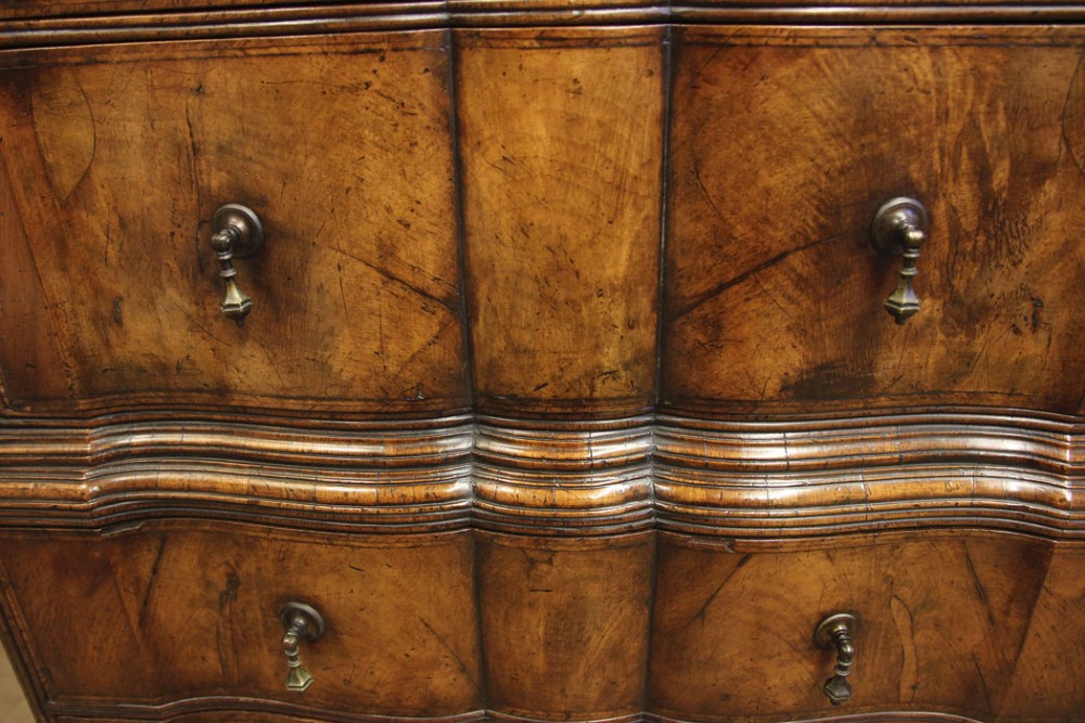 An antique figured walnut chest on chest featuring a serpentine shaped front, brass drop handles, and turned bun feet. View VI