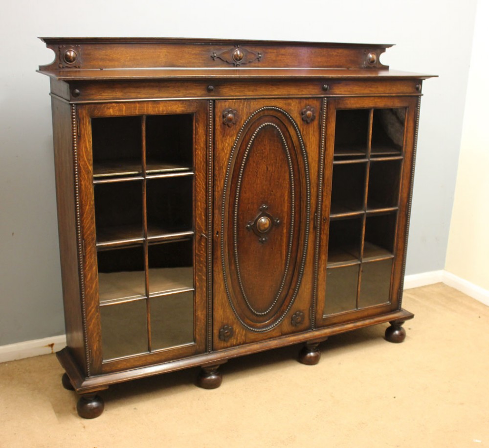 An antique dark oak low bookcase featuring twin glazed outer doors, a central carved oval timber door, and heavy bun feet.