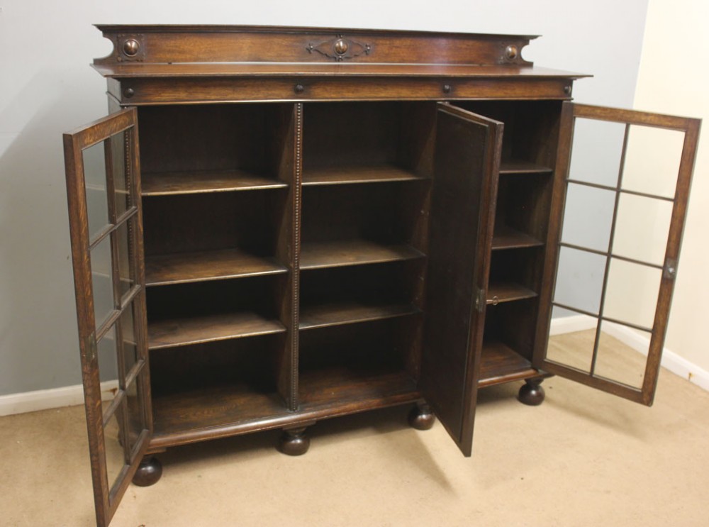 An antique dark oak low bookcase featuring twin glazed outer doors, a central carved oval timber door, and heavy bun feet. View II