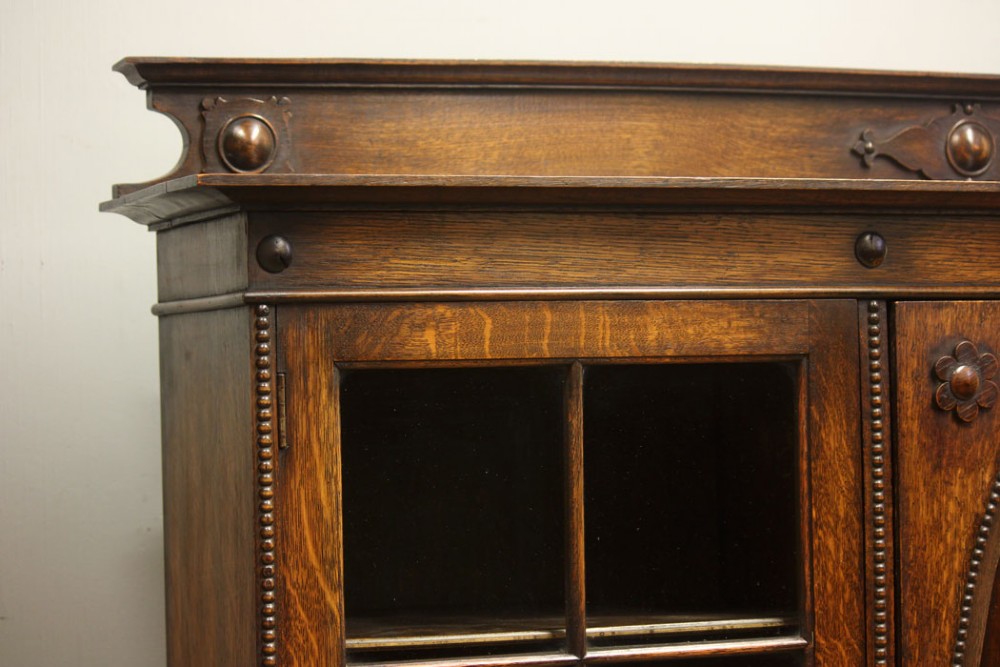 An antique dark oak low bookcase featuring twin glazed outer doors, a central carved oval timber door, and heavy bun feet. View V