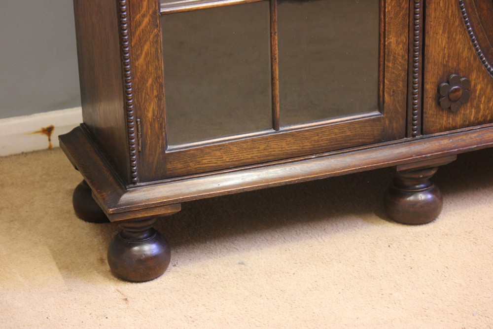 An antique dark oak low bookcase featuring twin glazed outer doors, a central carved oval timber door, and heavy bun feet. View VI