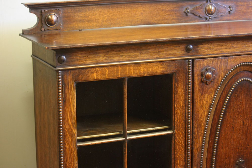 An antique dark oak low bookcase featuring twin glazed outer doors, a central carved oval timber door, and heavy bun feet. View VII