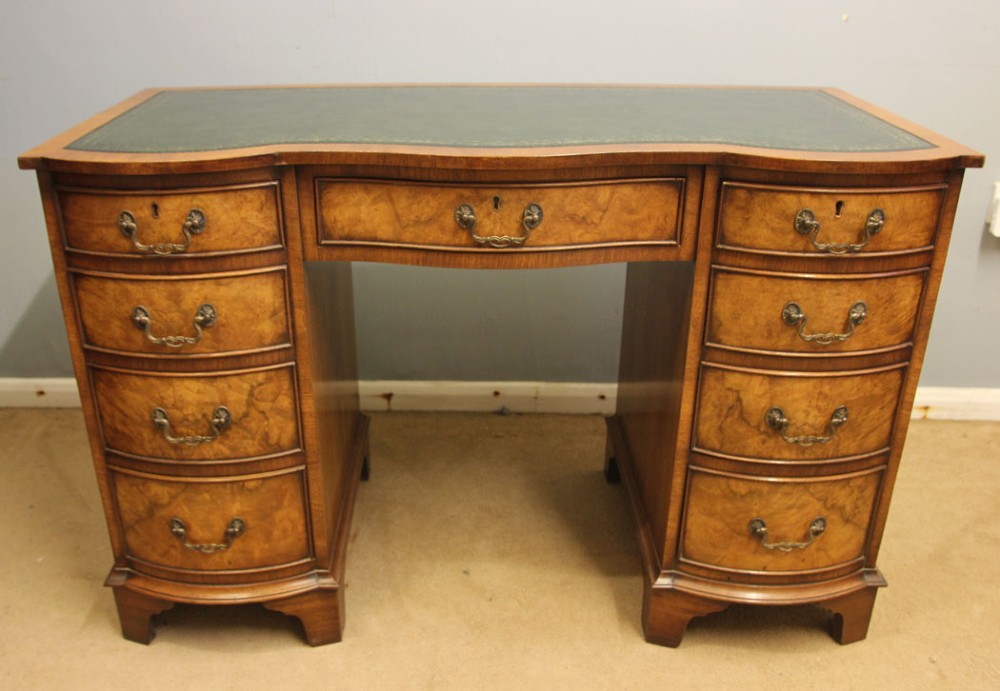 An antique burr walnut kneehole pedestal writing desk featuring a green leather inset top, nine drawers, and decorative brass handles.