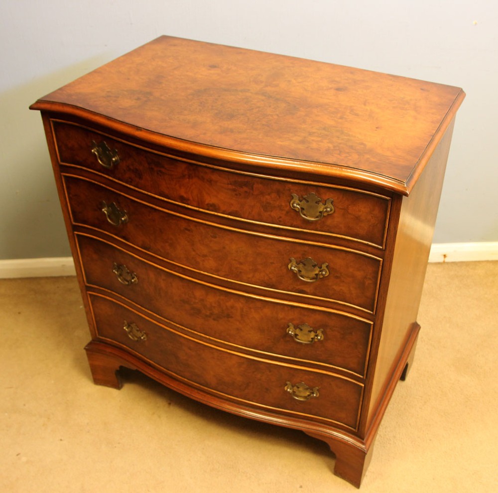 A burr walnut chest of drawers featuring four graduated drawers with a serpentine curved front, decorative brass handles, and resting on bracket feet. View VI