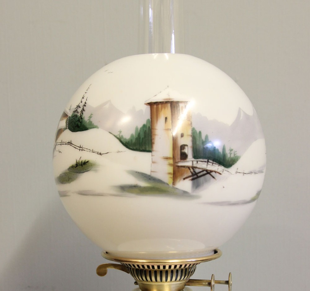 An antique oil lamp featuring a stepped brass Corinthian column base, a white glass font, a clear glass chimney, and a spherical white glass shade painted with a winter landscape. View V