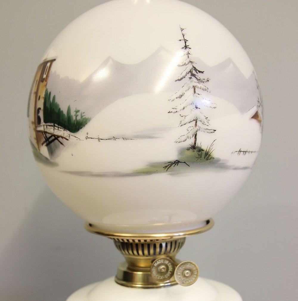 An antique oil lamp featuring a stepped brass Corinthian column base, a white glass font, a clear glass chimney, and a spherical white glass shade painted with a winter landscape. View VII