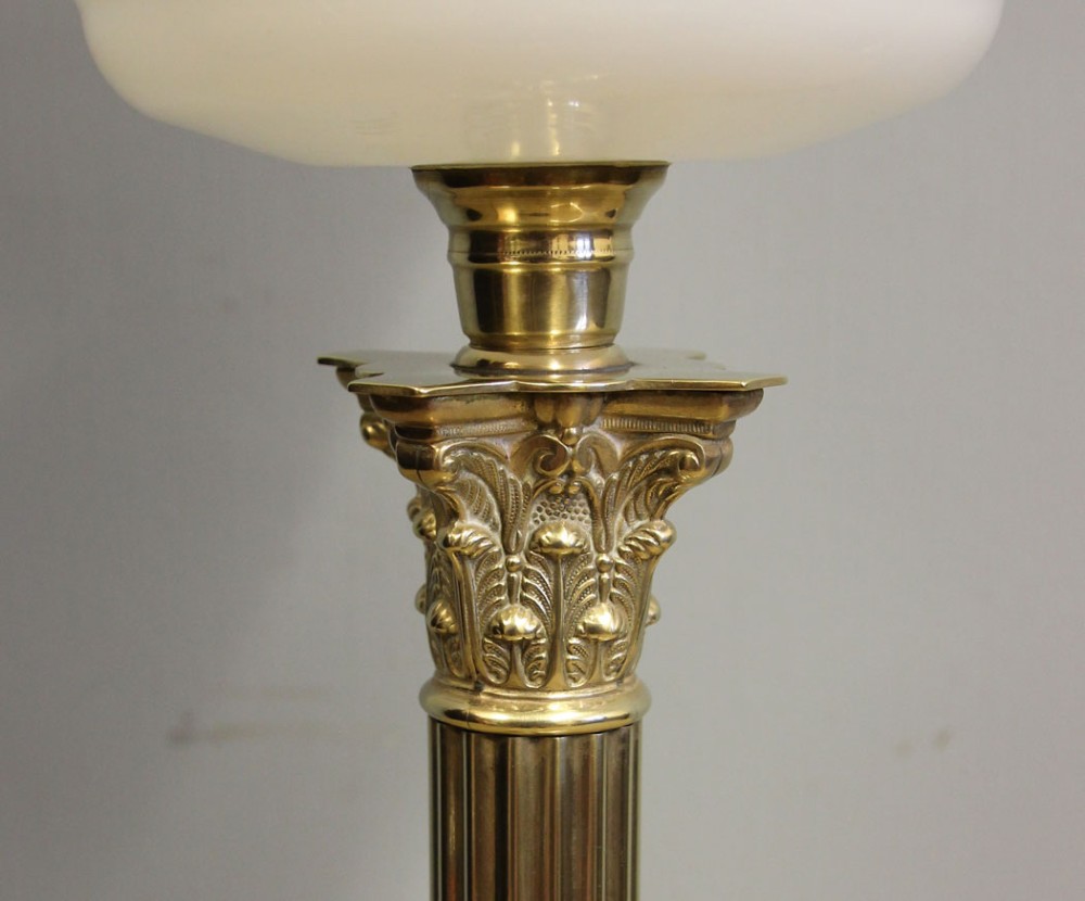 An antique oil lamp featuring a stepped brass Corinthian column base, a white glass font, a clear glass chimney, and a spherical white glass shade painted with a winter landscape. View X
