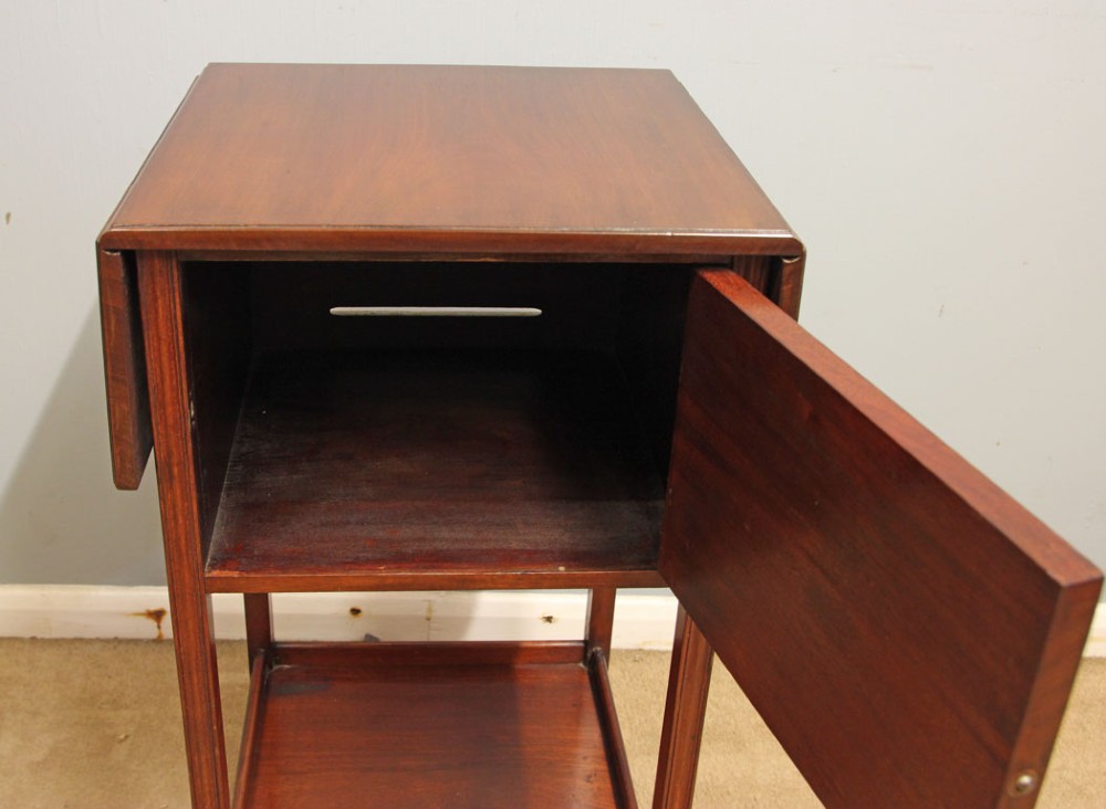 An antique mahogany bedside cabinet featuring two drop-leaf sides, a single door with a brass ring pull handle, straight legs, and an undertier shelf. View VI