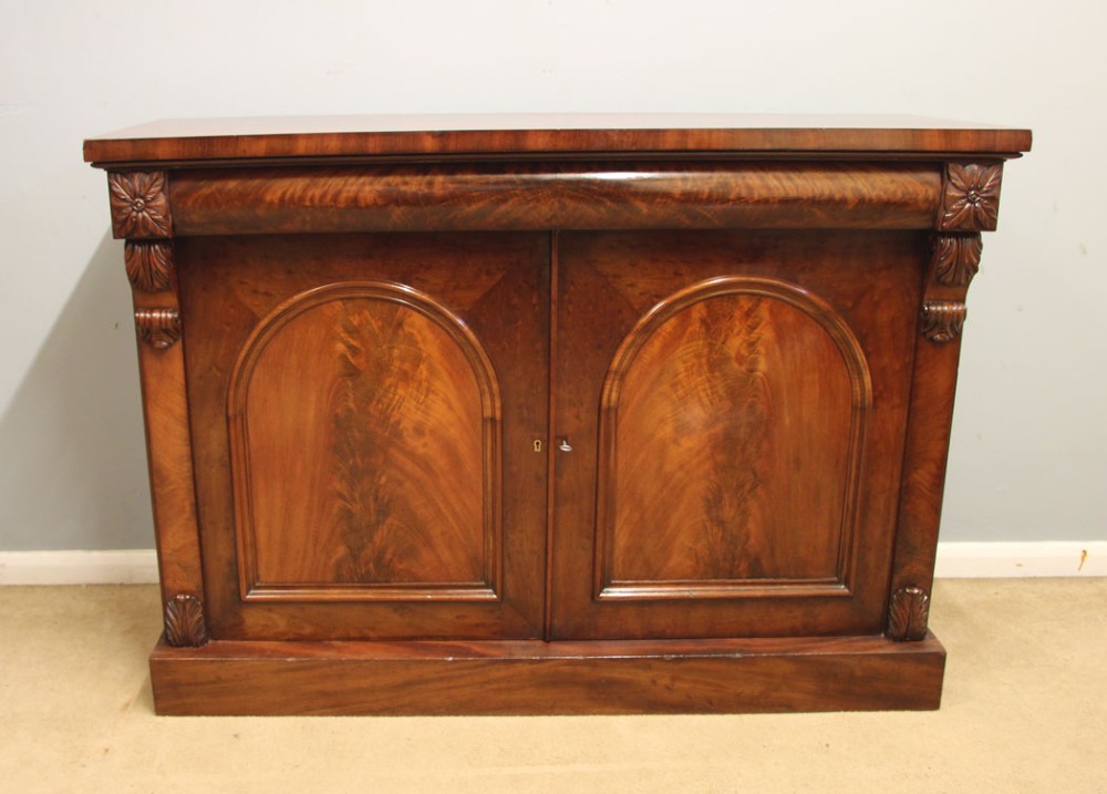 An antique Victorian flame mahogany chiffonier sideboard with two arched panel doors, carved corbels, and a plinth base.