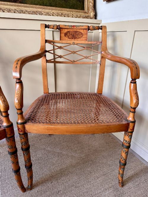 fig 1. A pair of Edwardian Sheraton Revival painted wooden elbow chairs featuring woven cane seats, lattice backrests, and floral painted decoration.