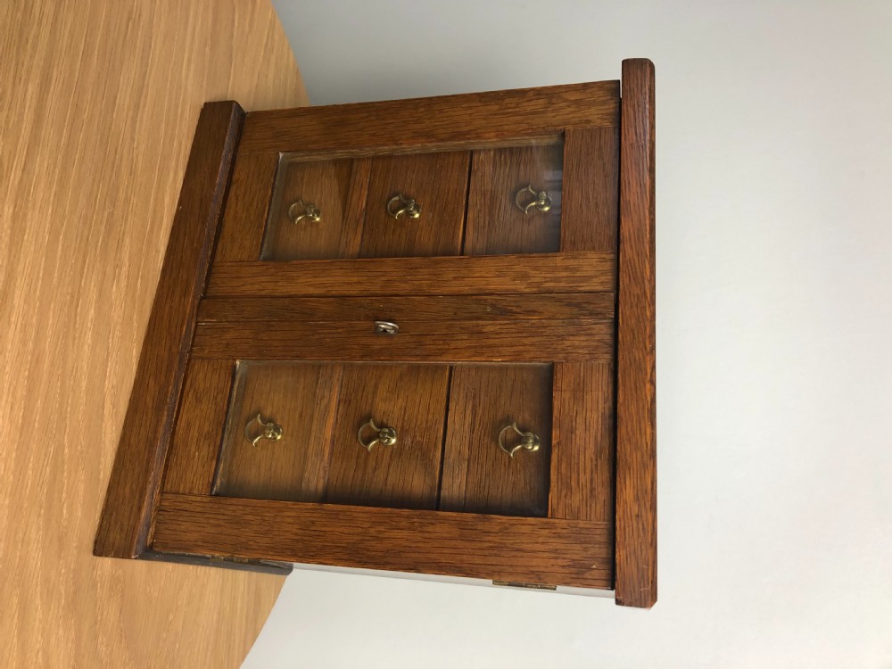 An Edwardian medium oak tabletop cabinet featuring twin glazed doors, six interior drawers with brass teardrop handles, and a central keyhole with key. View II