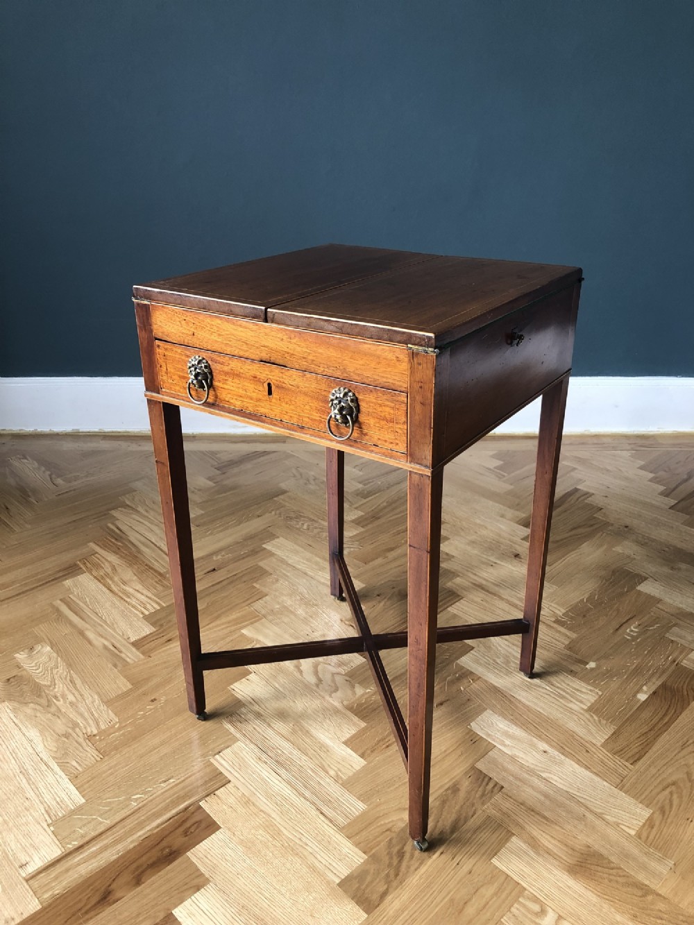 An antique Regency mahogany fold-out games table featuring a hinged top, a front drawer with brass lion-mask ring handles, tapered square legs, an X-stretcher base, and small castors.