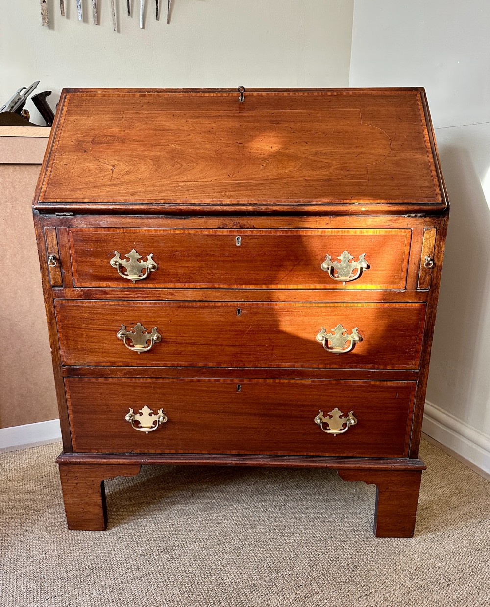 A small antique mahogany slope-front bureau featuring three drawers with brass batwing handles and keyholes, raised on bracket feet. View VI