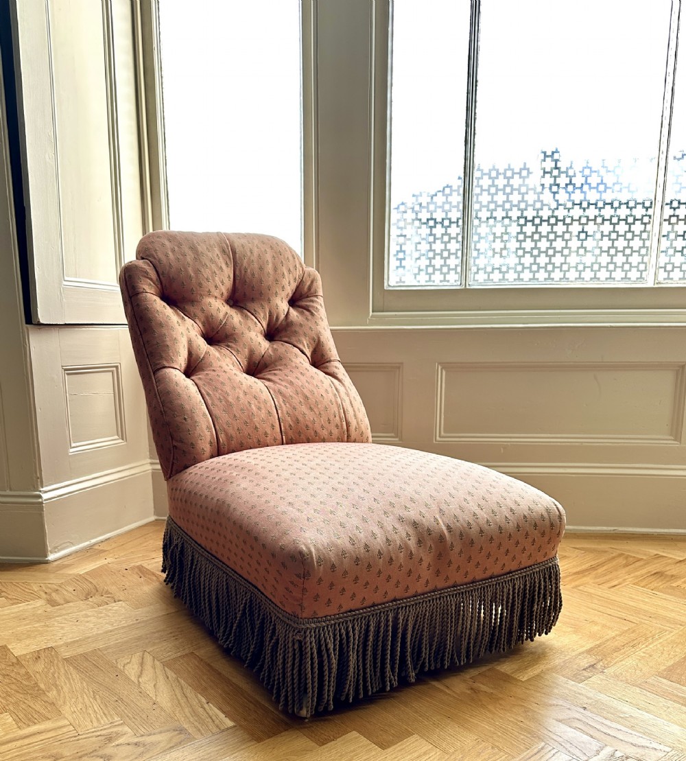 A 19th-century low armless slipper chair upholstered in a patterned dusty pink fabric featuring button tufting on the backrest and dense fringe along the base.