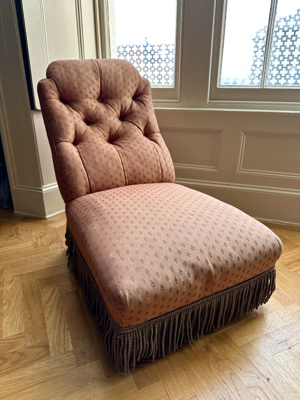 A 19th-century low armless slipper chair upholstered in a patterned dusty pink fabric featuring button tufting on the backrest and dense fringe along the base. View II