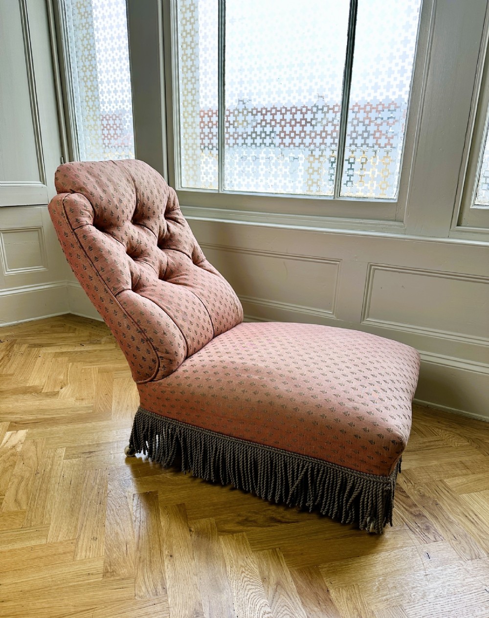 A 19th-century low armless slipper chair upholstered in a patterned dusty pink fabric featuring button tufting on the backrest and dense fringe along the base. View V