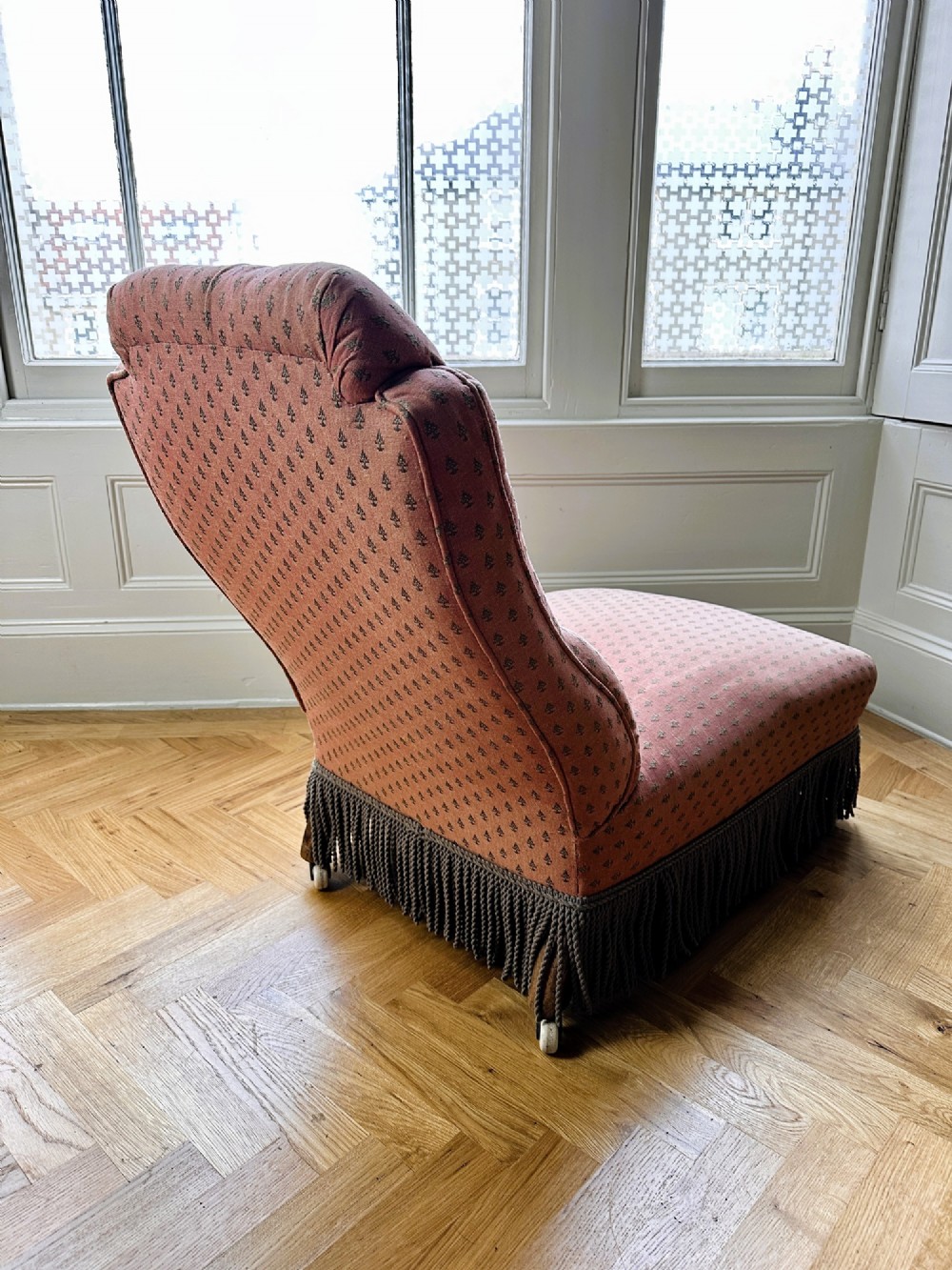 A 19th-century low armless slipper chair upholstered in a patterned dusty pink fabric featuring button tufting on the backrest and dense fringe along the base. View VI