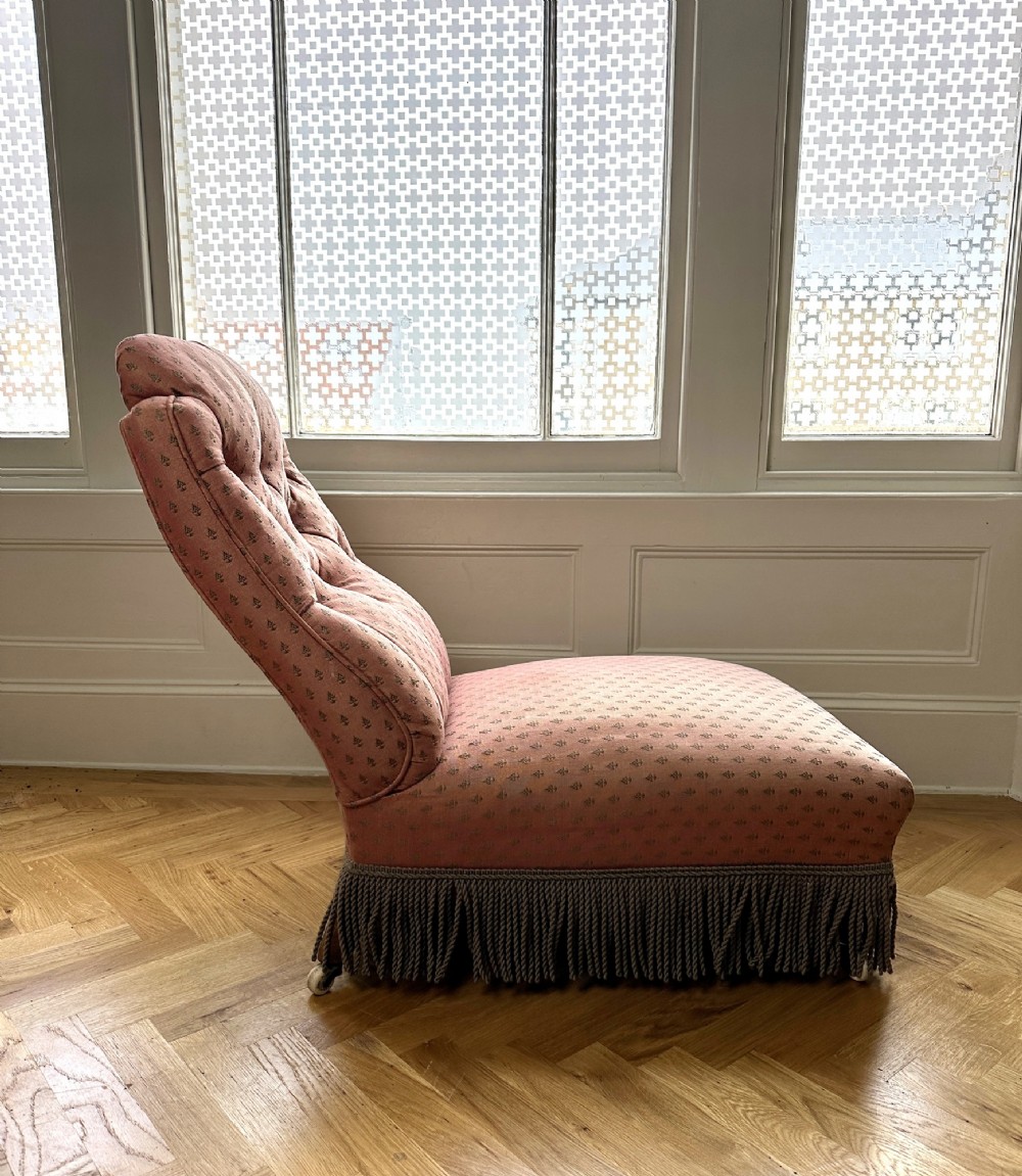 A 19th-century low armless slipper chair upholstered in a patterned dusty pink fabric featuring button tufting on the backrest and dense fringe along the base. View VII