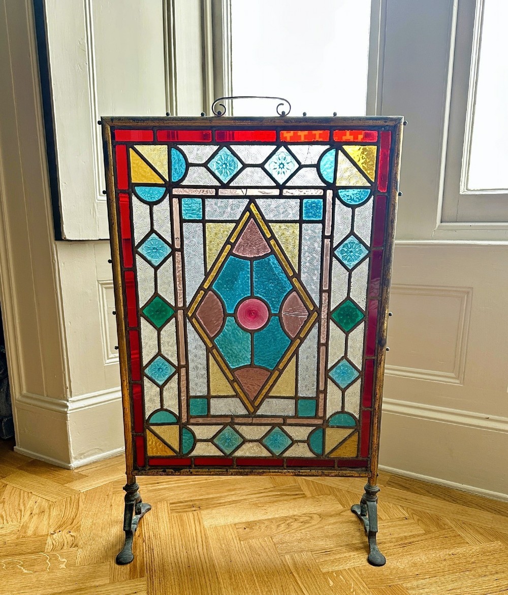 An Arts and Crafts period firescreen featuring a geometric leaded stained glass panel in red, blue, green, and yellow hues set within an iron frame.