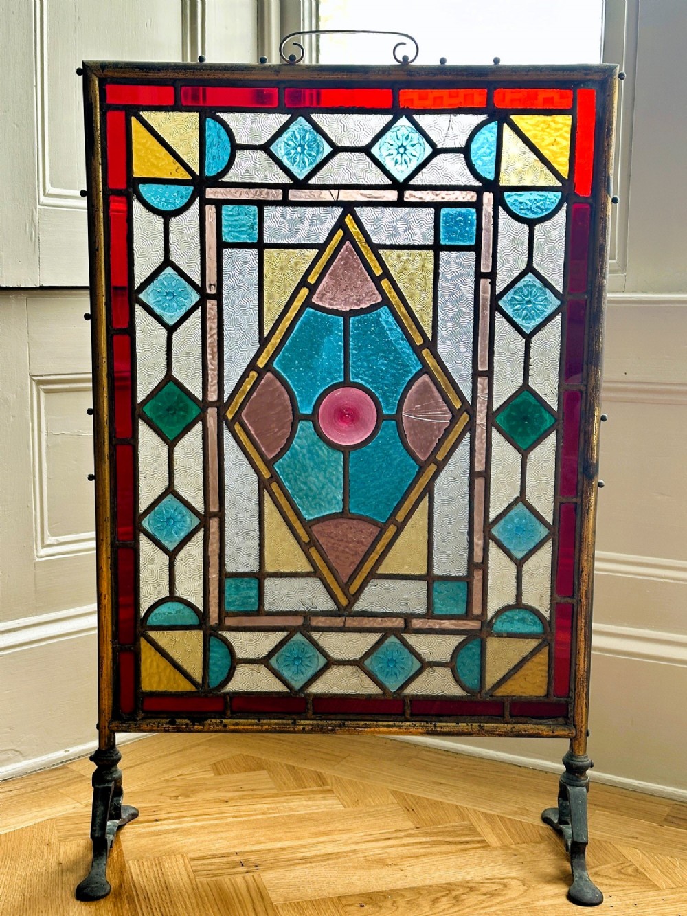 An Arts and Crafts period firescreen featuring a geometric leaded stained glass panel in red, blue, green, and yellow hues set within an iron frame. View II