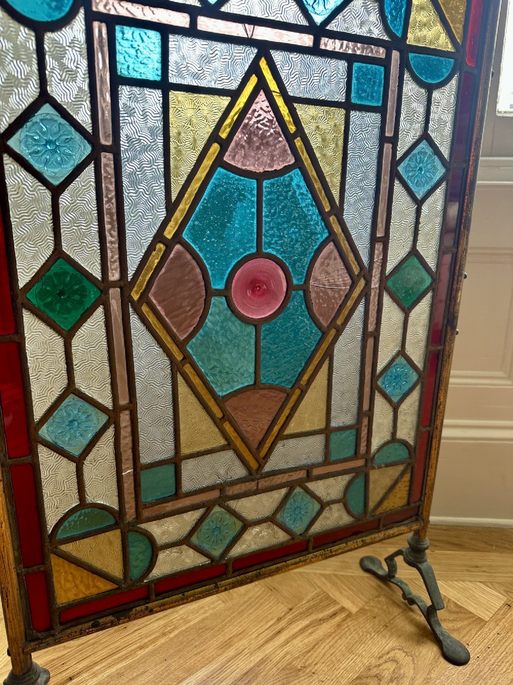 An Arts and Crafts period firescreen featuring a geometric leaded stained glass panel in red, blue, green, and yellow hues set within an iron frame. View III