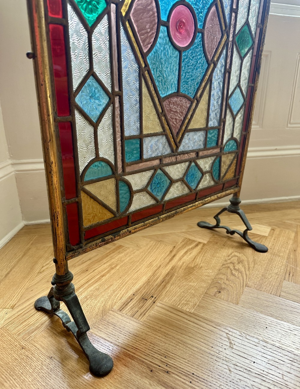 An Arts and Crafts period firescreen featuring a geometric leaded stained glass panel in red, blue, green, and yellow hues set within an iron frame. View IV