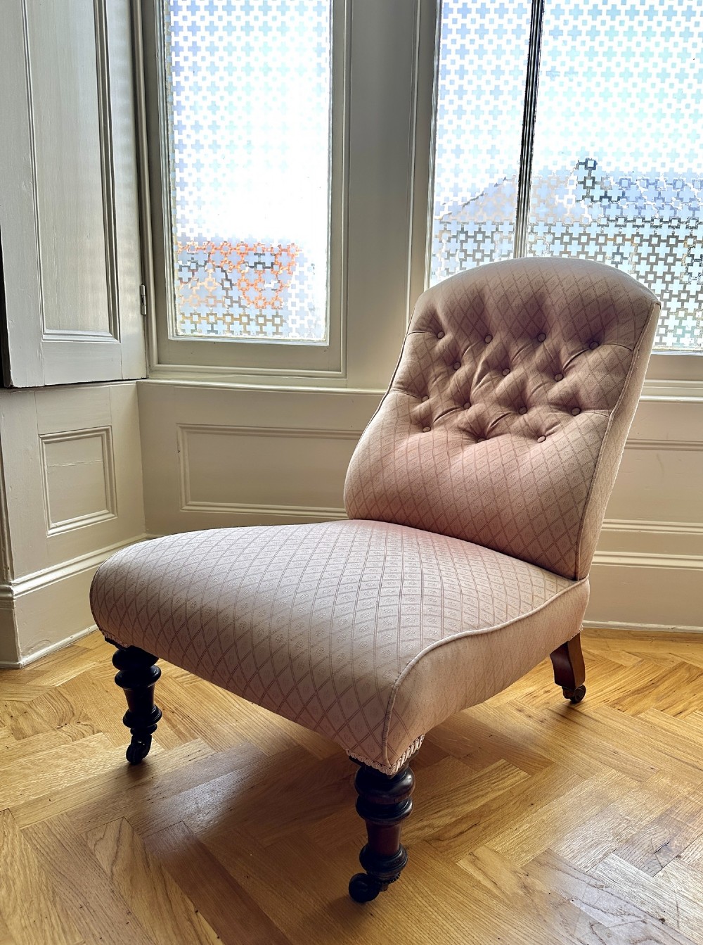 A low-seated Victorian slipper chair upholstered in dusty pink patterned fabric featuring a button-back design and turned dark timber legs with brass castors.