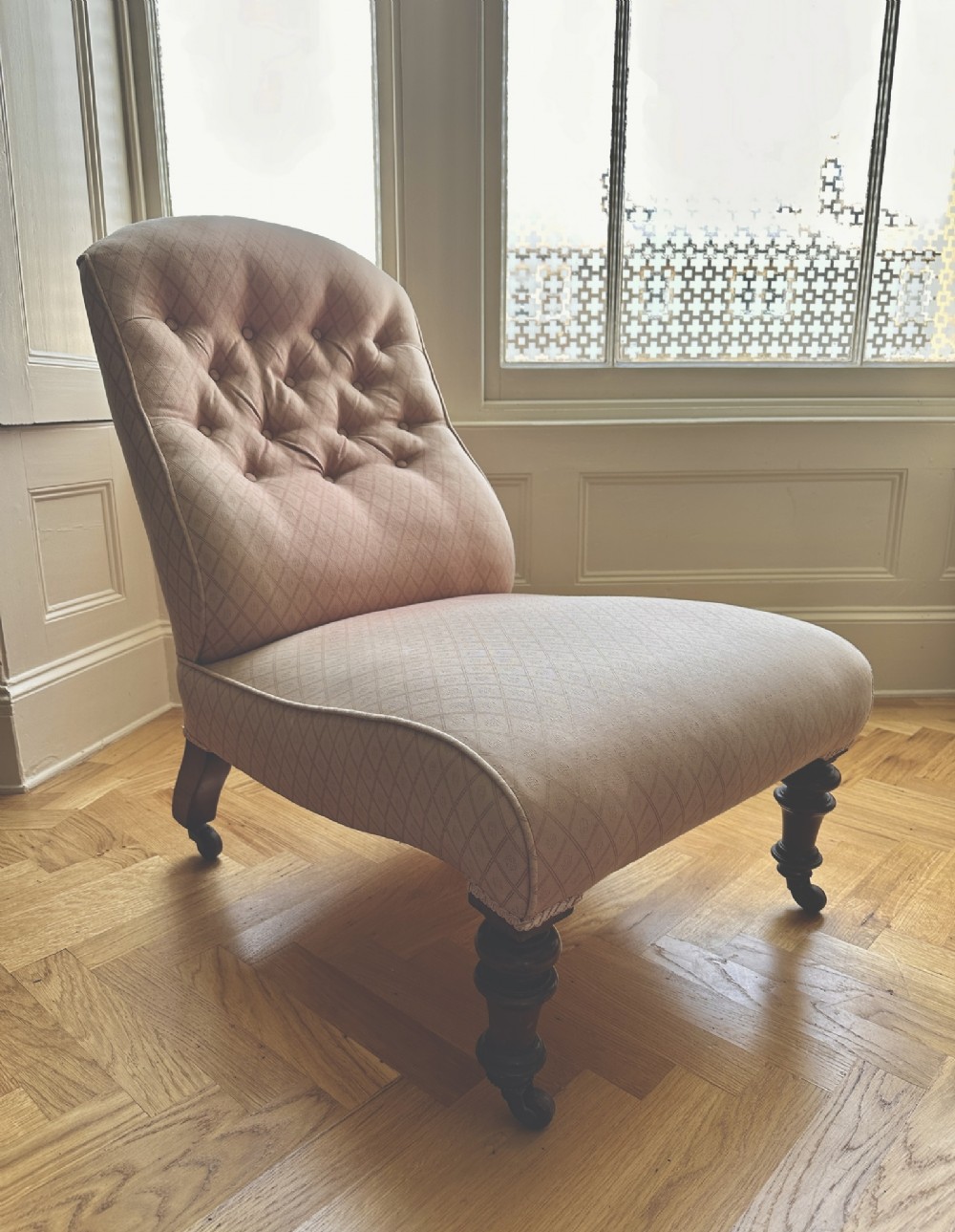 A low-seated Victorian slipper chair upholstered in dusty pink patterned fabric featuring a button-back design and turned dark timber legs with brass castors. View II