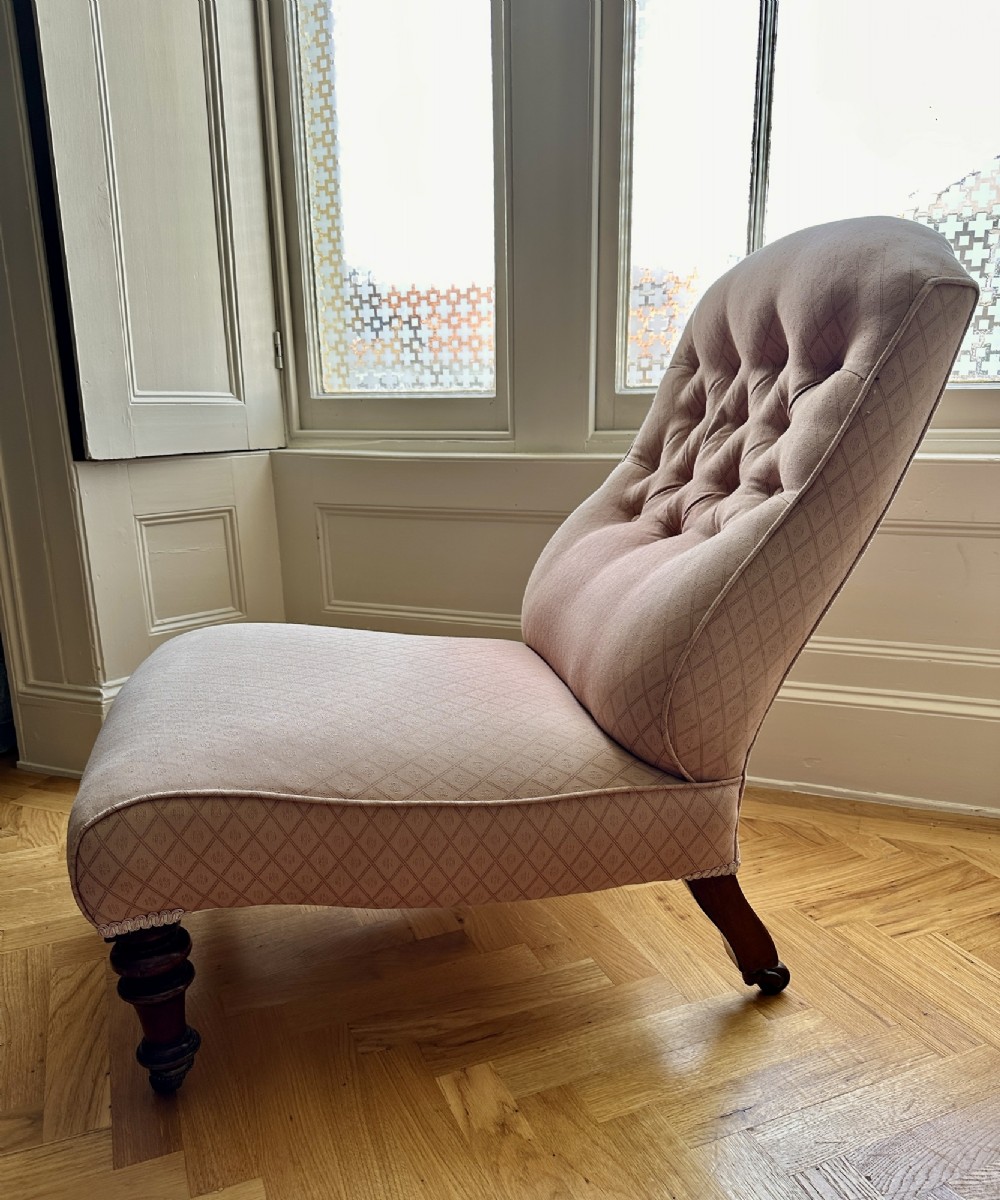 A low-seated Victorian slipper chair upholstered in dusty pink patterned fabric featuring a button-back design and turned dark timber legs with brass castors. View V