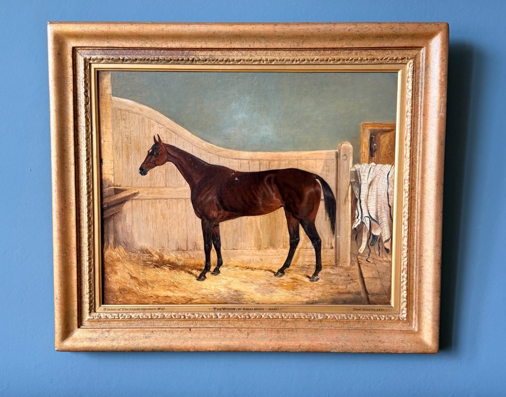 A Victorian oil painting depicting a brown racehorse standing in a straw-filled stable stall within a gilded frame.