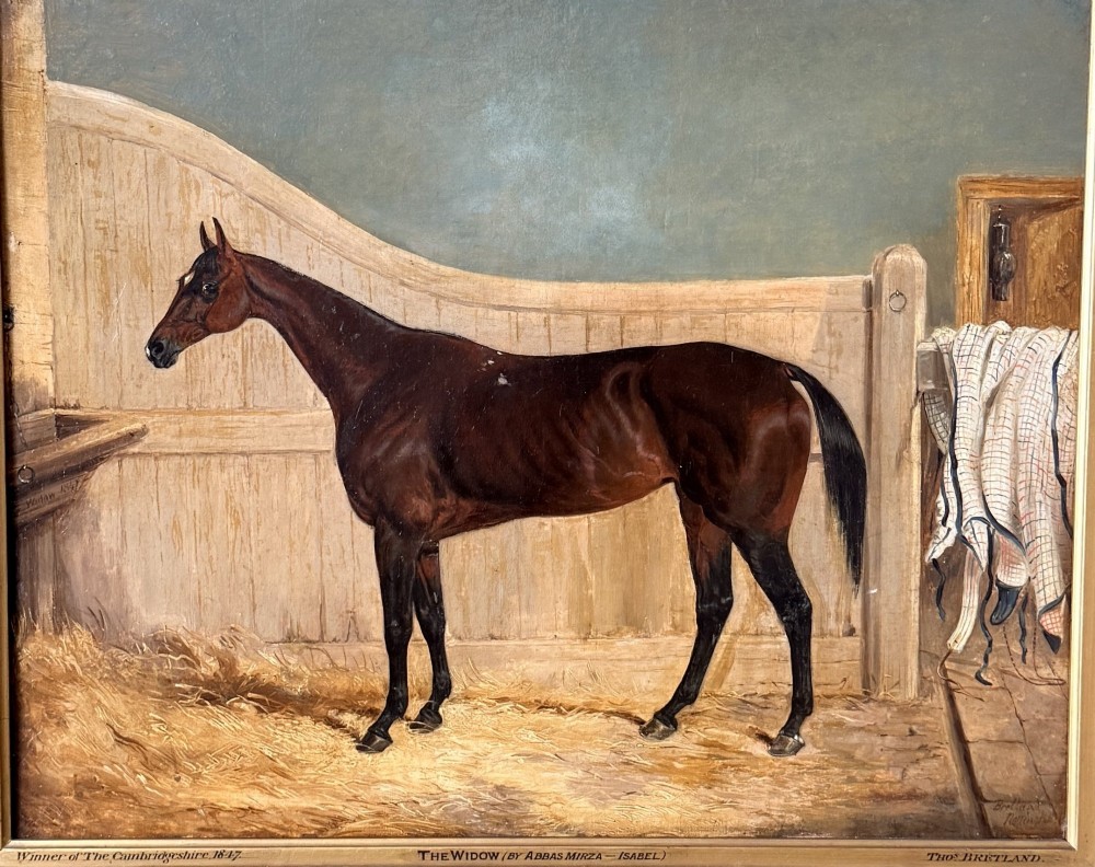 A Victorian oil painting depicting a brown racehorse standing in a straw-filled stable stall within a gilded frame. View II