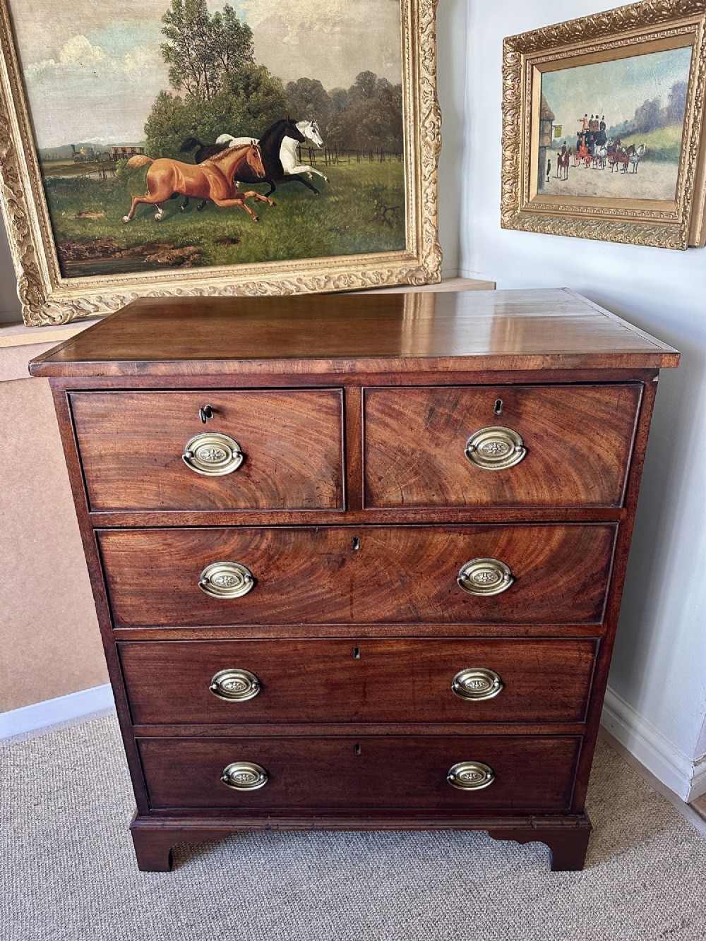 A late Georgian flame mahogany chest of drawers featuring a two-over-three drawer configuration, brass oval handles, bracket feet, and a polished wood finish.