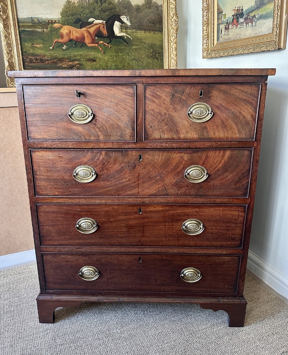 A late Georgian flame mahogany chest of drawers featuring a two-over-three drawer configuration, brass oval handles, bracket feet, and a polished wood finish. View II
