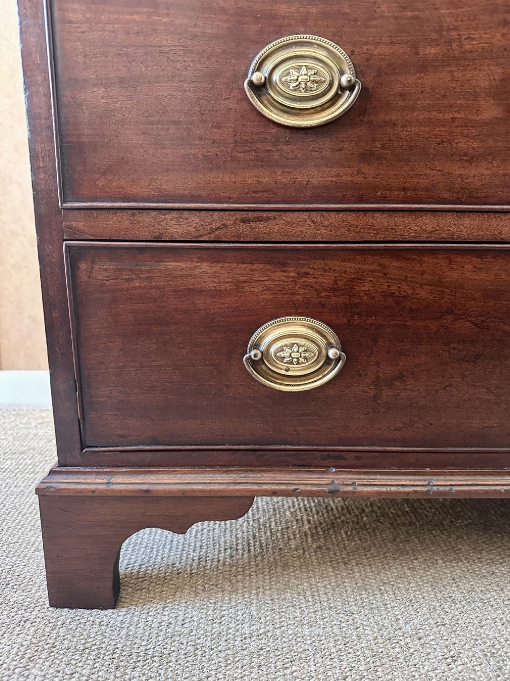 A late Georgian flame mahogany chest of drawers featuring a two-over-three drawer configuration, brass oval handles, bracket feet, and a polished wood finish. View V