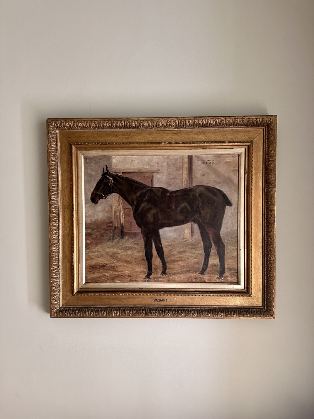 An 1896 oil painting depicting a dark brown horse standing inside a stable, housed within a gilded frame with a brass nameplate.