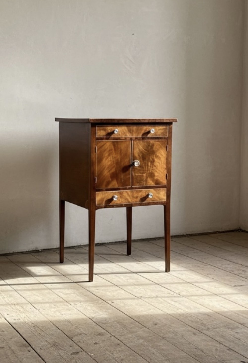 A Georgian flame mahogany bedside cabinet featuring a top drawer, central double doors, and a bottom drawer, supported by square tapered legs.