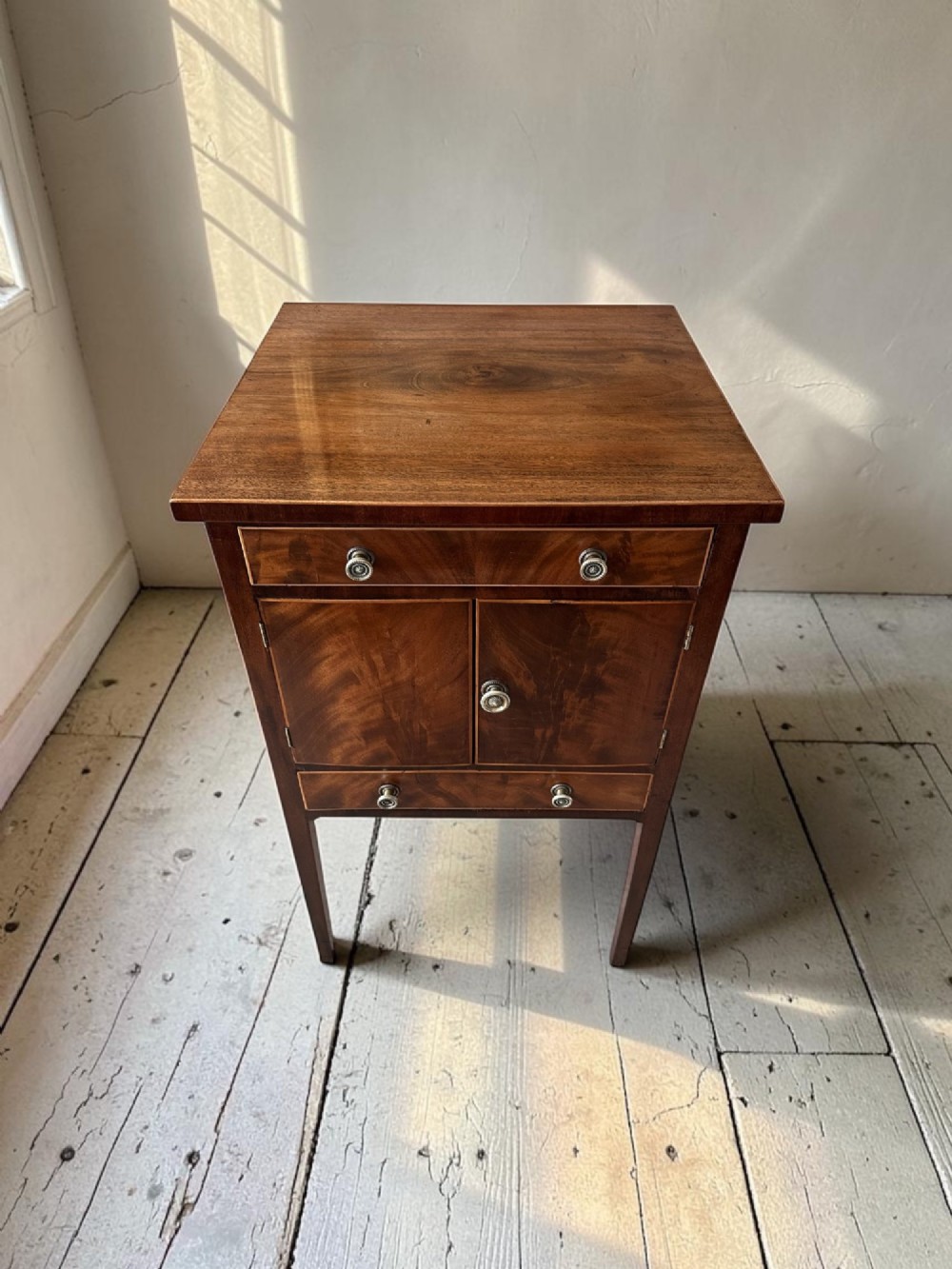 A Georgian flame mahogany bedside cabinet featuring a top drawer, central double doors, and a bottom drawer, supported by square tapered legs. View III
