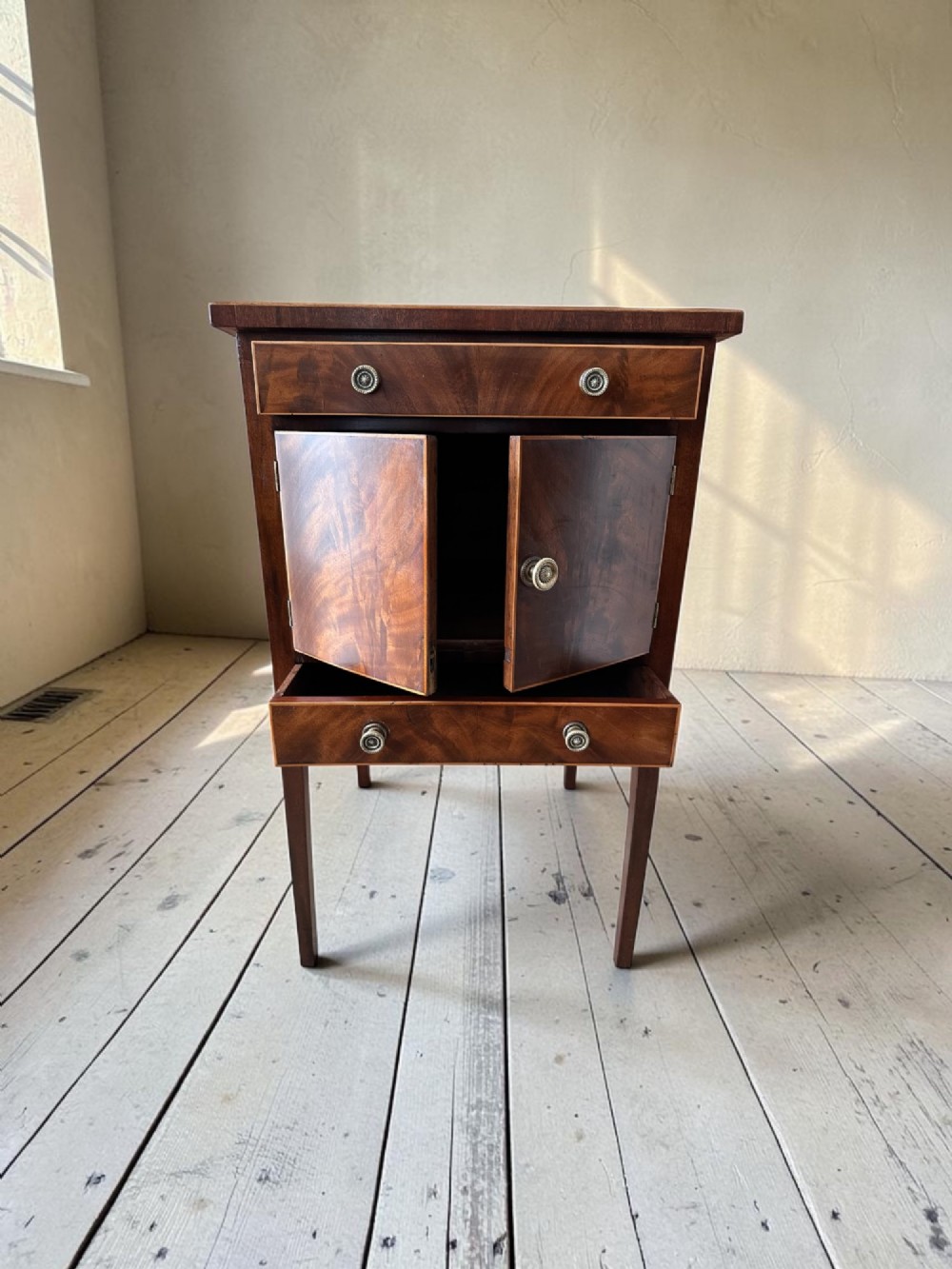 A Georgian flame mahogany bedside cabinet featuring a top drawer, central double doors, and a bottom drawer, supported by square tapered legs. View IV