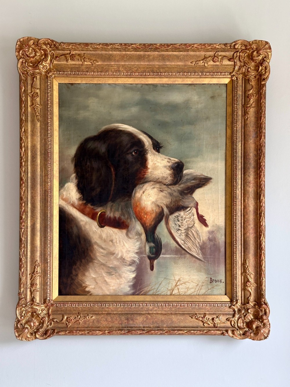 An early 20th-century oil painting depicting a brown and white gundog holding a mallard drake in its mouth, presented within an ornate gilt frame.