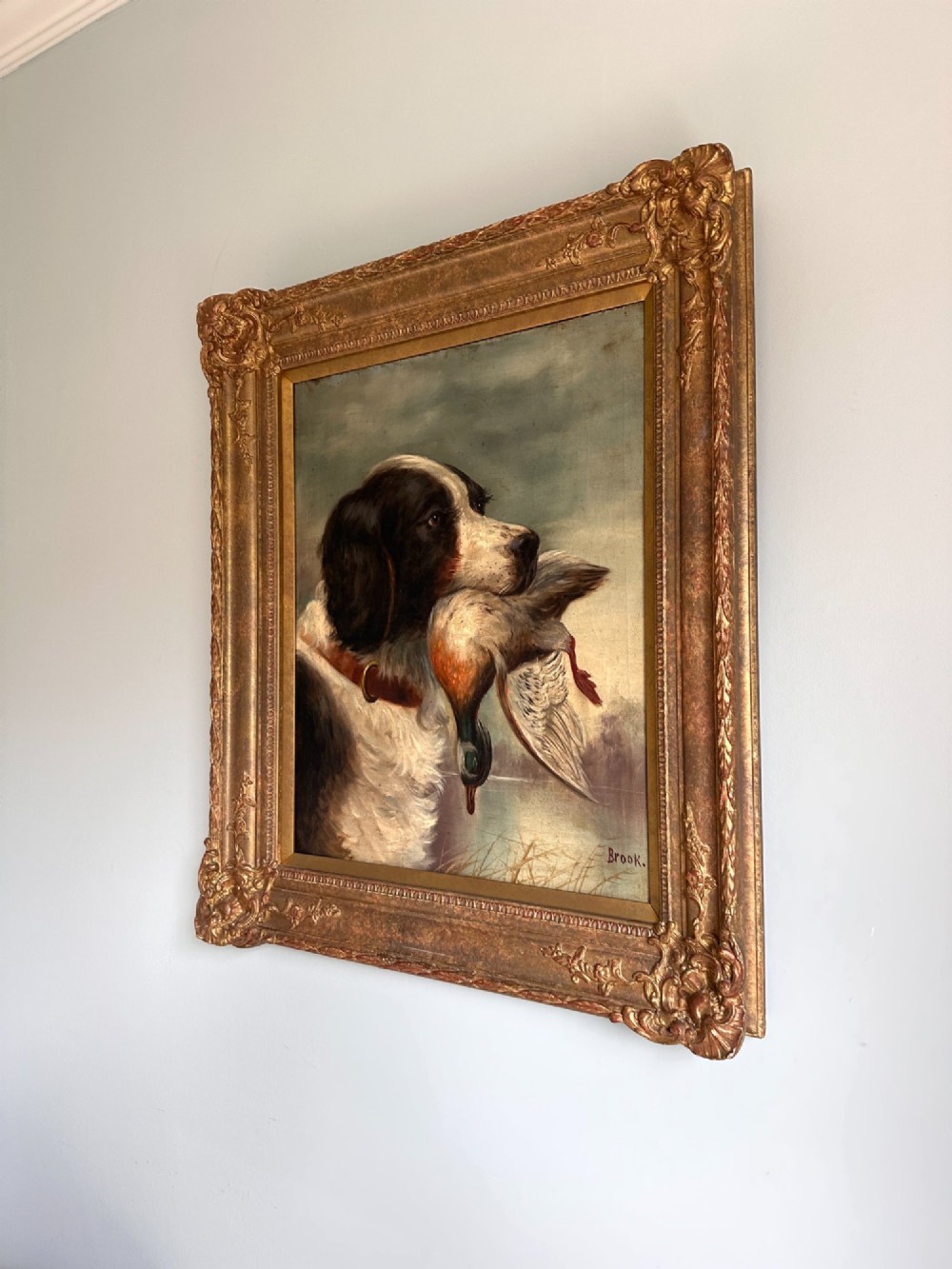 An early 20th-century oil painting depicting a brown and white gundog holding a mallard drake in its mouth, presented within an ornate gilt frame. View V