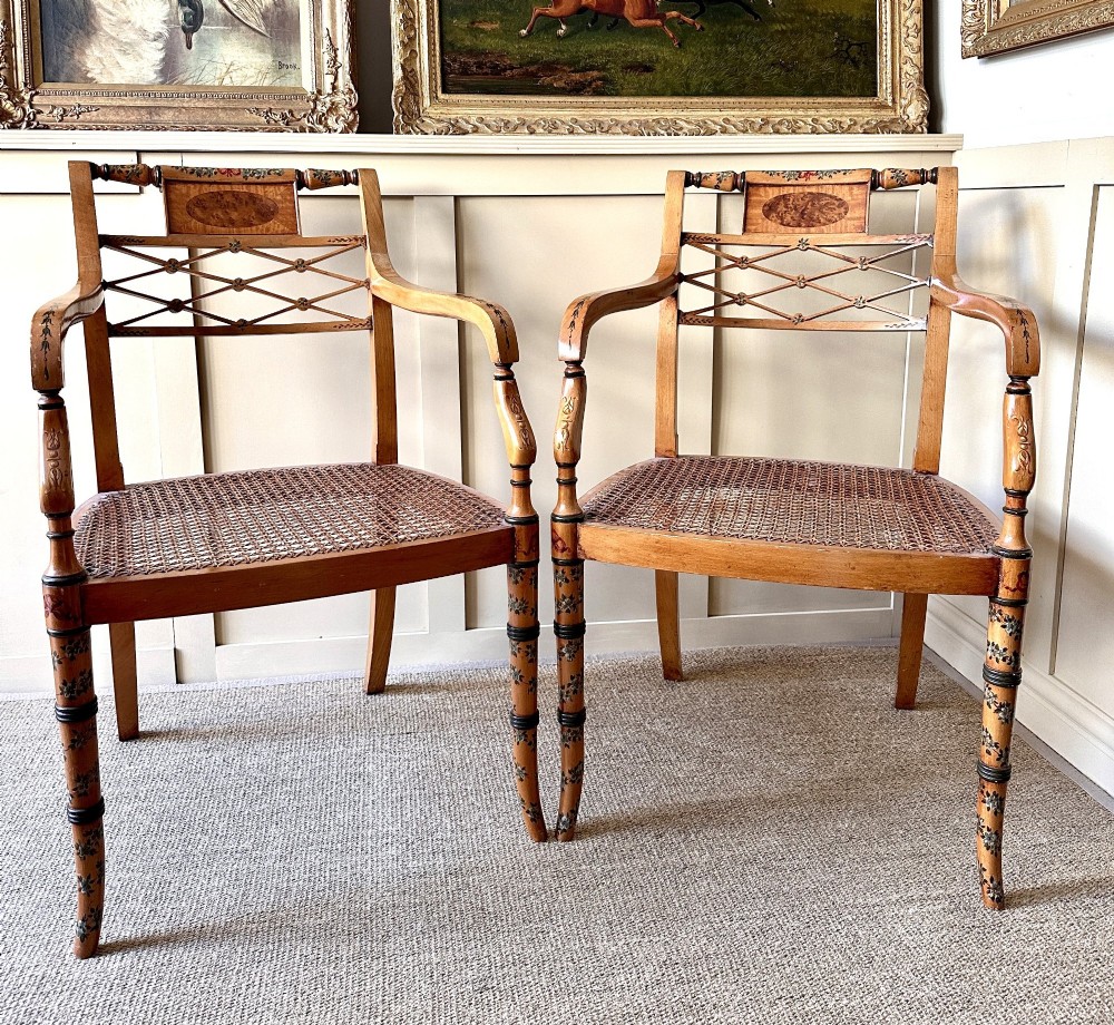 A pair of Edwardian Sheraton Revival painted wooden elbow chairs featuring woven cane seats, lattice backrests, and floral painted decoration.
