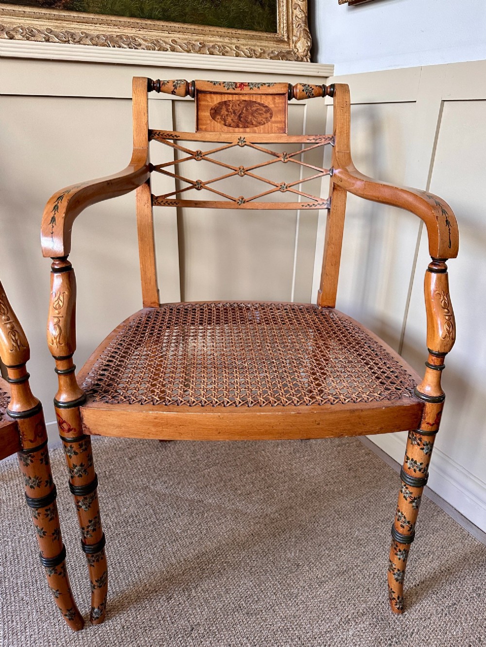 A pair of Edwardian Sheraton Revival painted wooden elbow chairs featuring woven cane seats, lattice backrests, and floral painted decoration. View II