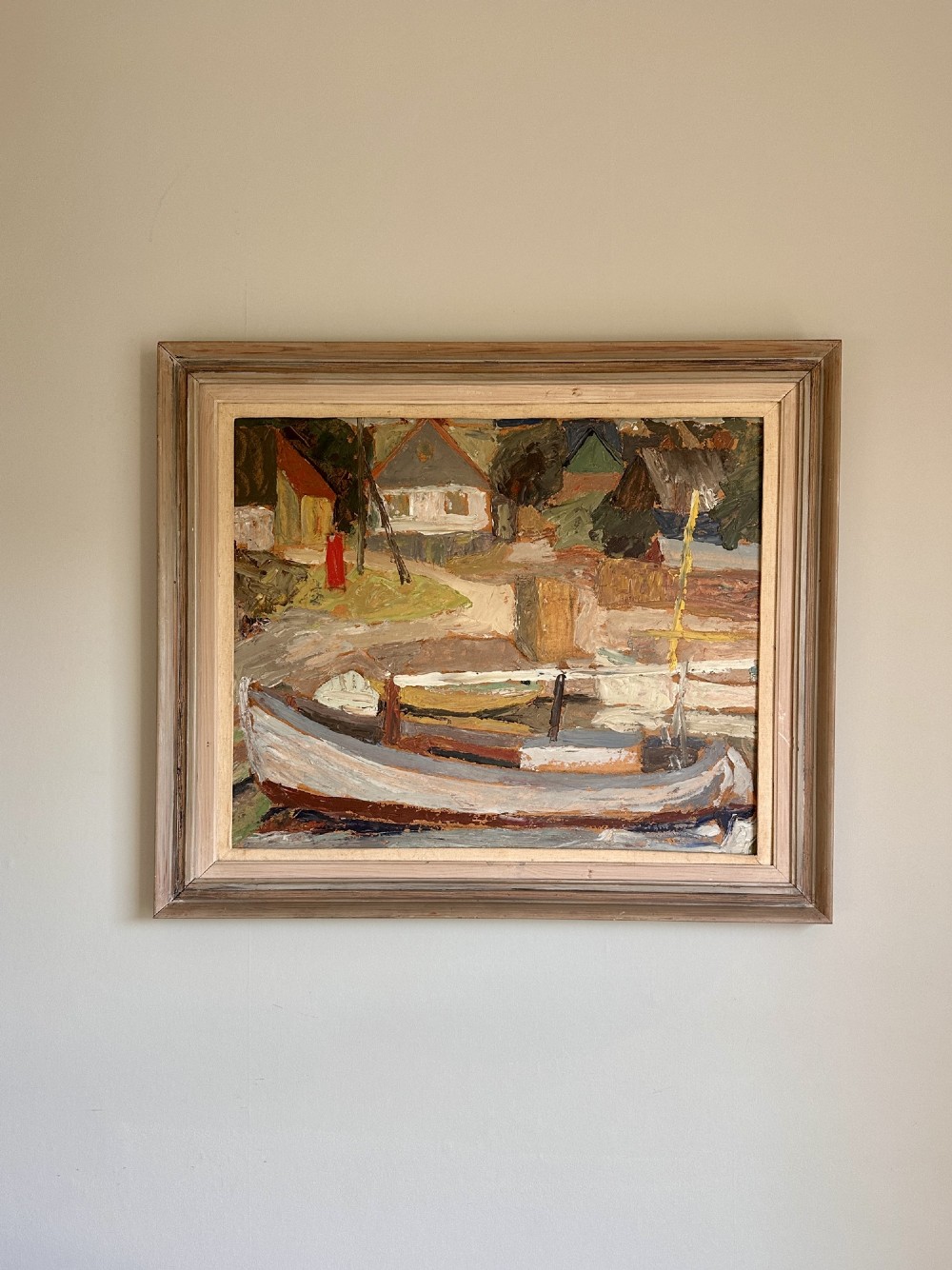 A framed mid-century oil painting depicting a harbour scene with grounded fishing boats and small coastal village houses rendered in textured impasto strokes and muted earth tones.