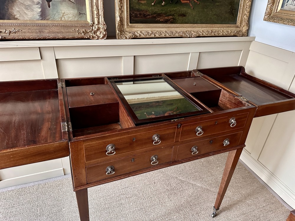 A late Georgian mahogany gentlemans dressing table featuring two hinged side leaves open to reveal internal compartments and a central adjustable mirror, raised on square tapered legs with brass castors. View VII