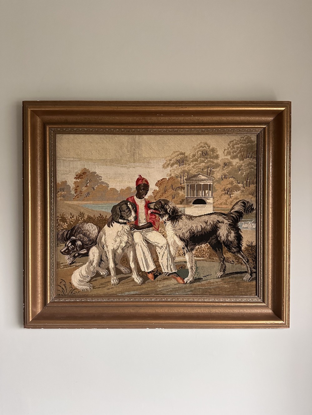 A framed Victorian Berlin woolwork tapestry depicting a seated figure with two dogs in a parkland setting within a moulded gilt frame.