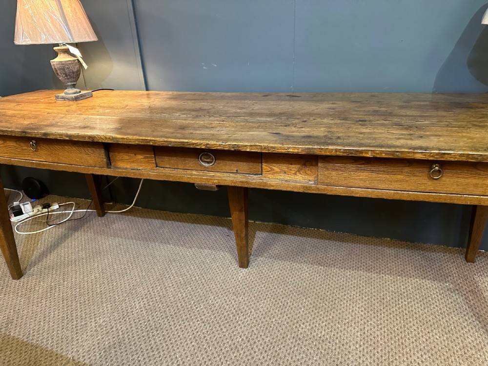 A long 19th-century oak table featuring a plank top, three front drawers with metal ring pulls, and four tapered square legs. View VI